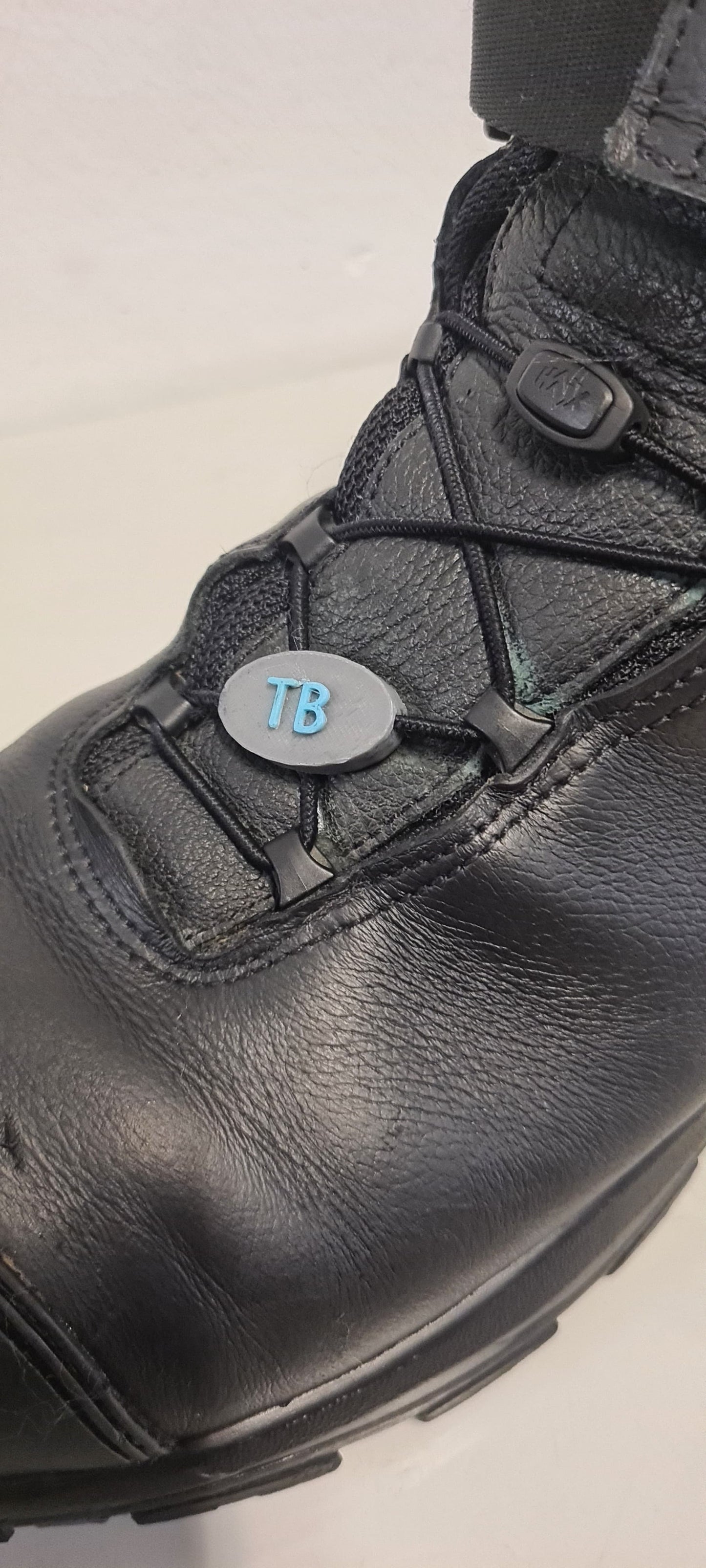 Ein schwarzer Haix Nevada Mid Stiefel mit personalisierbarem Namensschild aus PLA, das an den Schnürsenkeln befestigt ist. Das ovale Schild trägt die blauen Initialen "TB" und passt sich perfekt in das Schnellschnürsystem ein. Ideal für eine individuelle Kennzeichnung im Einsatz.