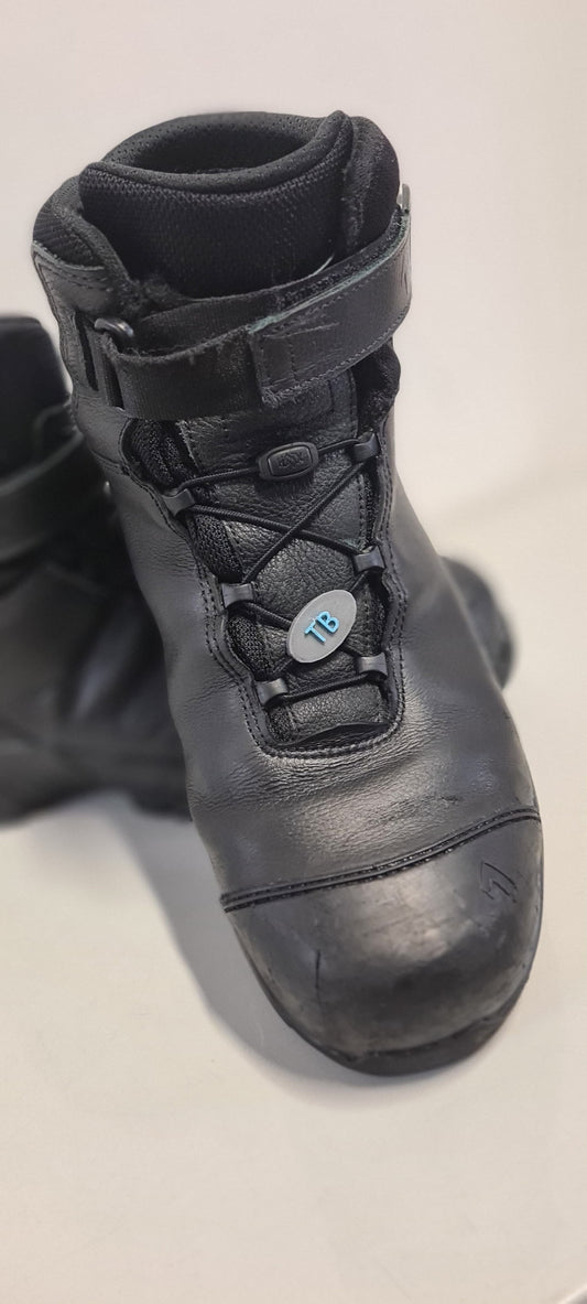 Ein schwarzer Haix Nevada Mid Stiefel mit personalisierbarem Namensschild aus PLA, das an den Schnürsenkeln befestigt ist. Das ovale Schild trägt die blauen Initialen "TB" und passt sich perfekt in das Schnellschnürsystem ein. Ideal für eine individuelle Kennzeichnung im Einsatz.