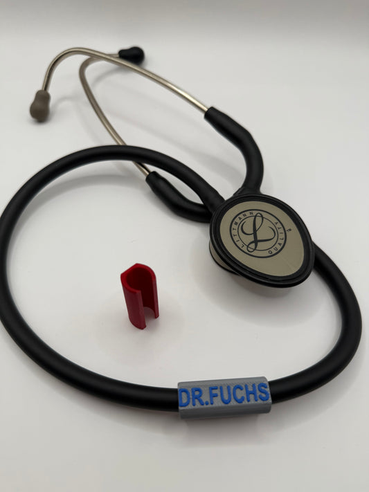 Hier sieht man ein Stethoskop, welches ein namensschild um seinen Schlauch hat, welches die Farbe in Grau hat, die Gravur oben drauf ist Dr.Fuchs in der Farbe Blau